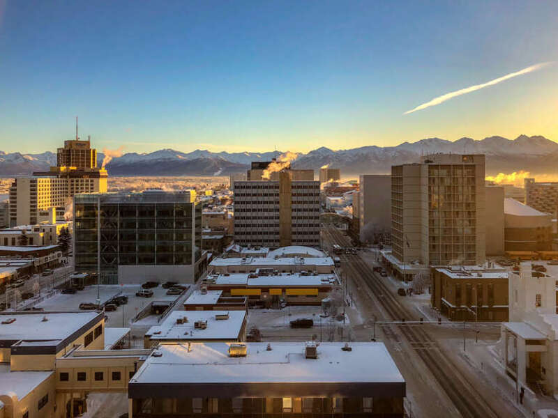 The sun sets on Downtown Anchorage on a cold, quiet day in February, with the Chugach Mountain Range just below the horizon above snow-covered rooftops, as seen from the Hotel Captain Cook.
