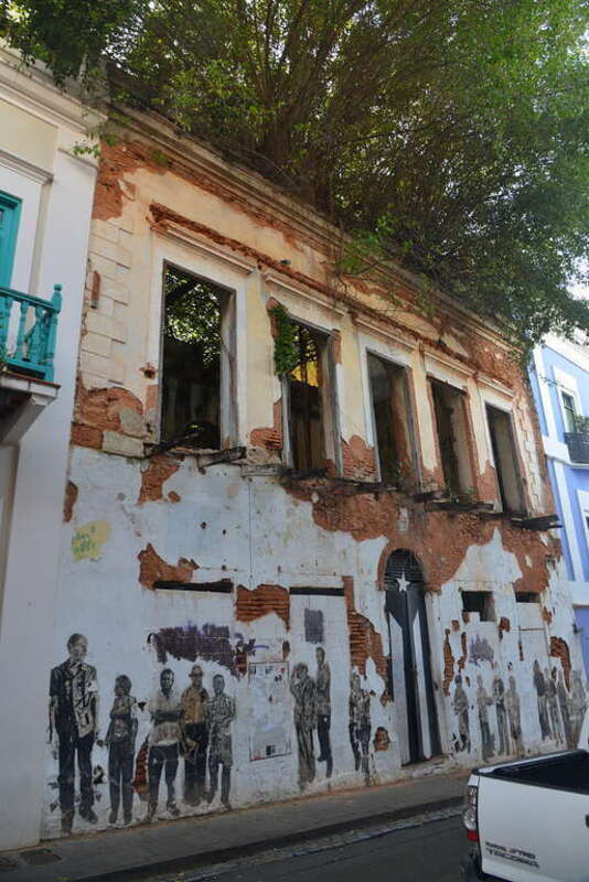 This building is just a shell with no roof and some large trees growing in it.  The front of the building has black and white depictions of people on it and the door, which used to be painted like the PUerto Rico flag in full color now has a black