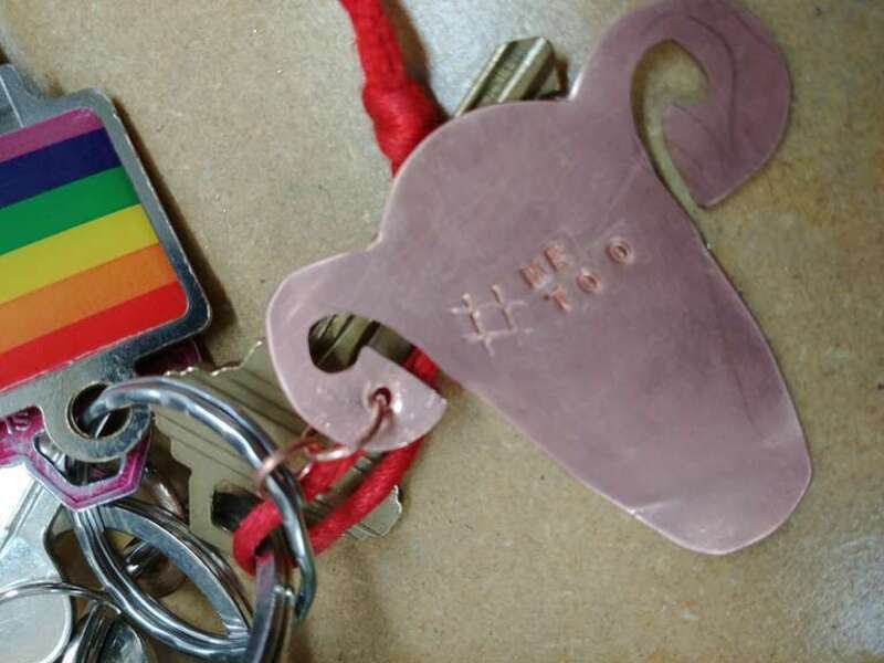 A keychain shaped like a uterus, stamped with &quot;#METOO&quot;. A key with a rainbow inset is also seen.