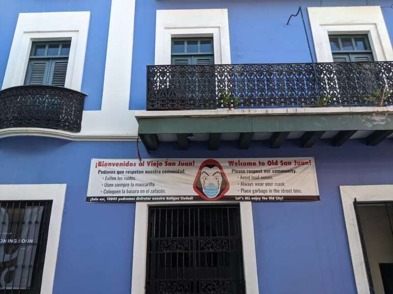 A banner on the front of a building in both English and Spanish that says: &quot;Welcome to Old San Juan! Please respect our community.  Avoid loud noises, Always wear your mask. Place garbage in the street bins. Let's ALL enjoy the Old City!&quot;