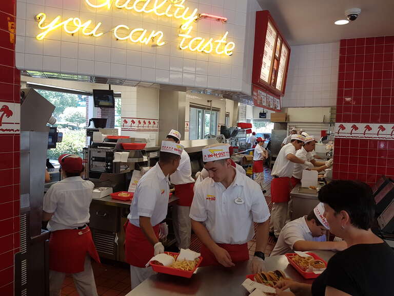 Standard kitchen and customer counter area of an In-n-Out branch. At high peak times up to 16 associates are working in abranch. One associate is outside registering drive-thru orders, another cleaning up in the dining area.