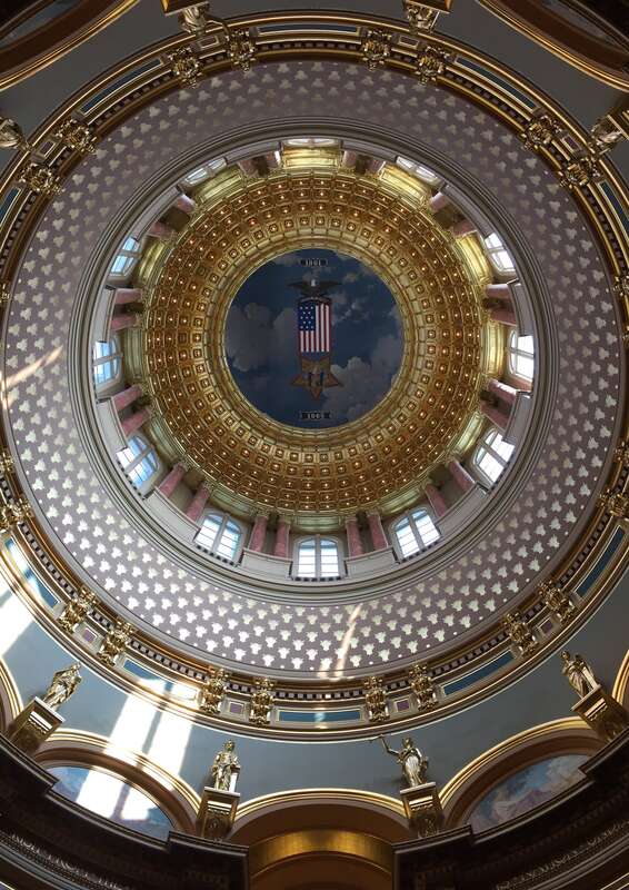 A photograph of the rotunda of the State Capitol of Iowa.  In the center suspended on wires beneath the painted ceiling is a large banner of the Grand Army of the Republic insignia.
