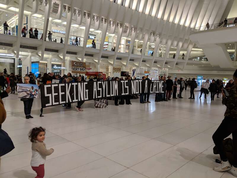 Photograph of members of the public carrying a large sign reading &quot;seeking asylum is not a crime&quot; in the Oculus building, in the World Trade Complex, in New York City.