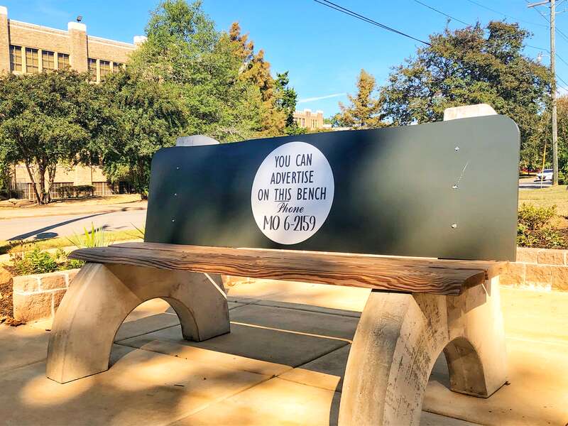 A replica bus bench with concrete curved legs, a wooden seat and black back rest with a circular advertisement reading &quot;You can advertise on this bench - phone MO 6-2159&quot; sits at the corner of 16th Street and S. Park Street. 
At this location,