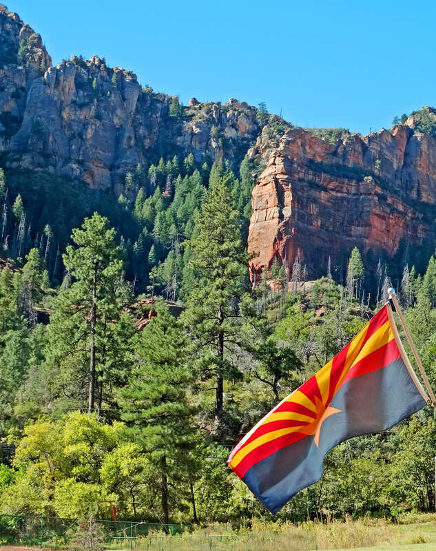 (1 in a multiple picture album)

Sitting in the shade of the general store I caught this image of the Arizona State Flag with the high, red-rock cliff and spires behind.