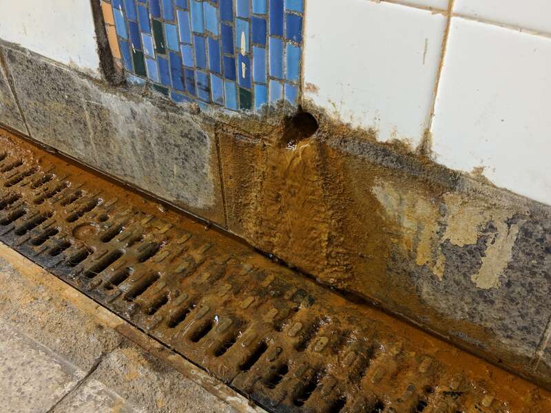 Hole in the wall in the New York City Subway producing rusted water, dripping into a dirty grate.