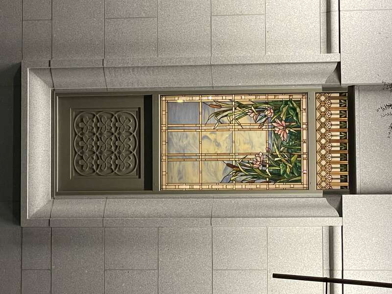 A stained glass window of the Orem Utah Temple, in the frame is a scene of bullrushes and cherry blossoms next to water. It is surrounded by a golden frame. Stone masonry can be seen off to the side, along with 9 cherry blossoms interlocked in a