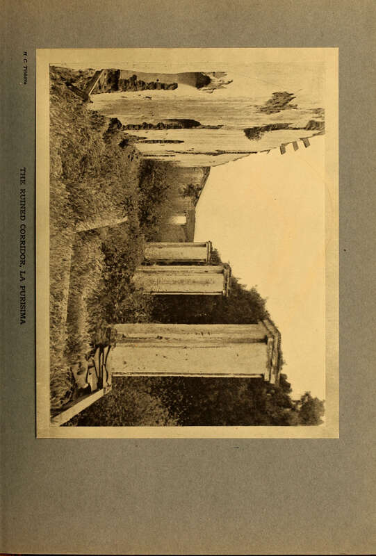Sepia toned photograph of the ruins of a un-roofed corridor over-grown with grass, columns to the right and wall with door to the left.