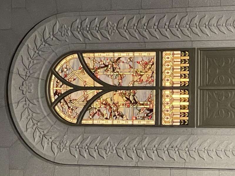 A stained glass window with branches depicting cherry blossoms and leaves, surrounded by gold. The window is surrounded by carved masonry, depicting branches and cherry blossoms stretching from the top of the window to the bottom along an arch.