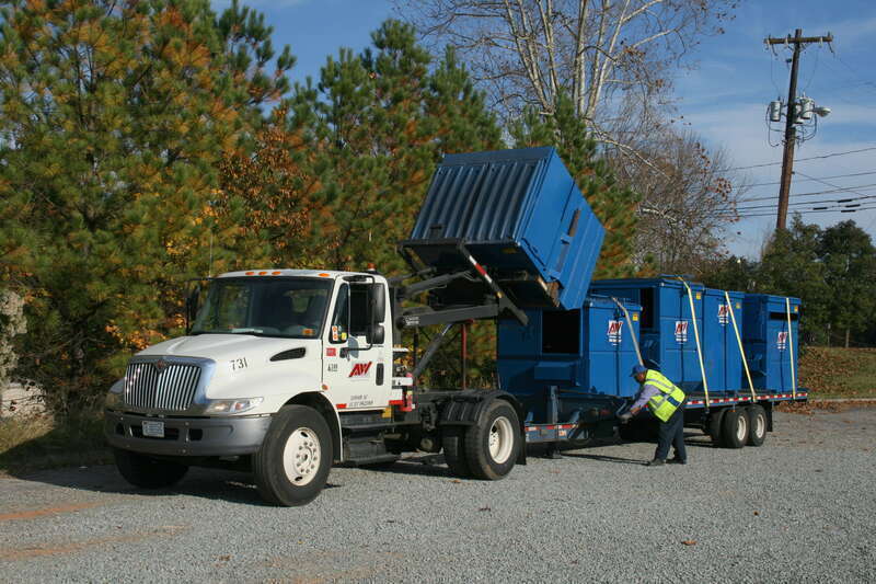 An operator unloading a blue Allied Waste Services dumpster from a specialised International truck onto a gravel lot at The Shoppes at Lakewood, a plaza in Durham, North Carolina.