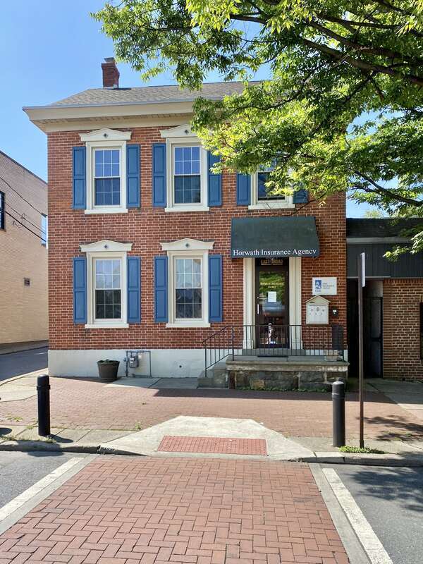 Built in the mid-19th Century, this Italianate-style three-bay red brick townhouse features a side gable roof, decorative window headers and stone sills, replacement windows, a stucco-clad stone base, a rusticated stone stoop, and a decorative
