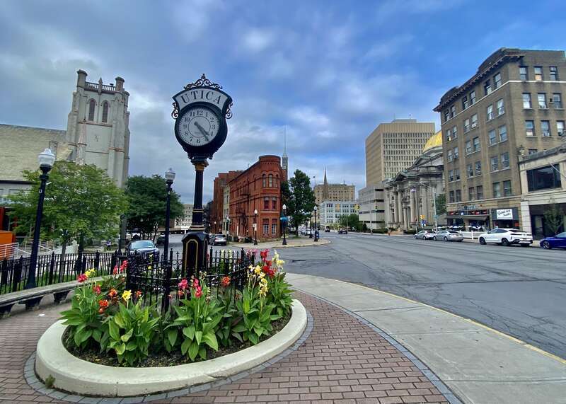 As seen in August 2021. One of the classic views of downtown Utica is the scene looking northeast along Genesee Street from the acute-angles corner of Washington Street, with the iconic vintage-style street clock in the left foreground and the