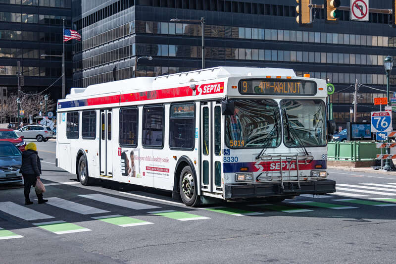 The SEPTA route 9 bus heading towards its terminus at 4th and Walnut. It is entering into the intersection crossing over the crosswalk and the dotted green bike lane crossing with the 22nd street trolley station entrance in the background as long as