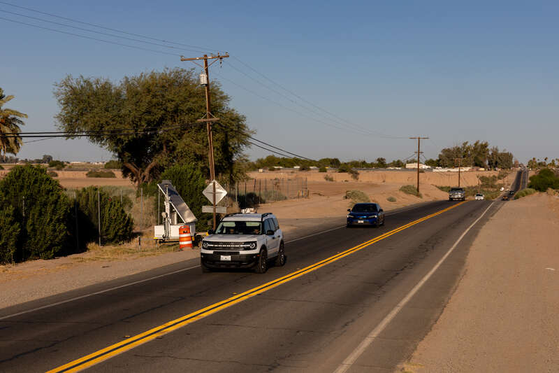 Two automated license plate reader (ALPR) systems—one disguised as a construction barrel and surveilling westbound traffic, the other held aloft in a metal box on the trailer beside it and surveilling eastbound traffic—on the north side of California