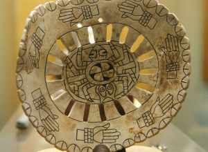 Spiro mound artifacts on display at Woolaroc Museum, near Bartlesville. Spider motif at center with circle of hands with shell bracelets. Roughly 3 in x 3 in.