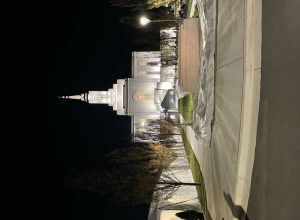 A picture of the Orem Utah Temple at night. Garden elements can be seen in the foreground, plastic wrapping shows underneath a sign that says: "The Church of Jesus Christ of Latter Day Saints, Orem Utah Temple."