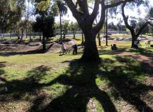 This is a midday photograph of the Inglewood Pumptrack from the north, facing south toward the bike facility. The Pumptrack's two tracks are visible in the picture. Just left of center is a looping track for riders to travel on BMX or mountain bikes.