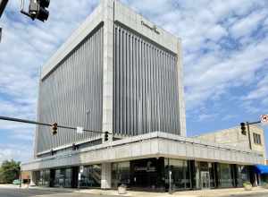 Built in 1969, this Modern International-style skyscraper features a stone panel and metal-clad exterior with a one-story podium featuring a large parapet and dark stone-clad columns, a plaza atop the roof of the podium with planters and flagpoles,